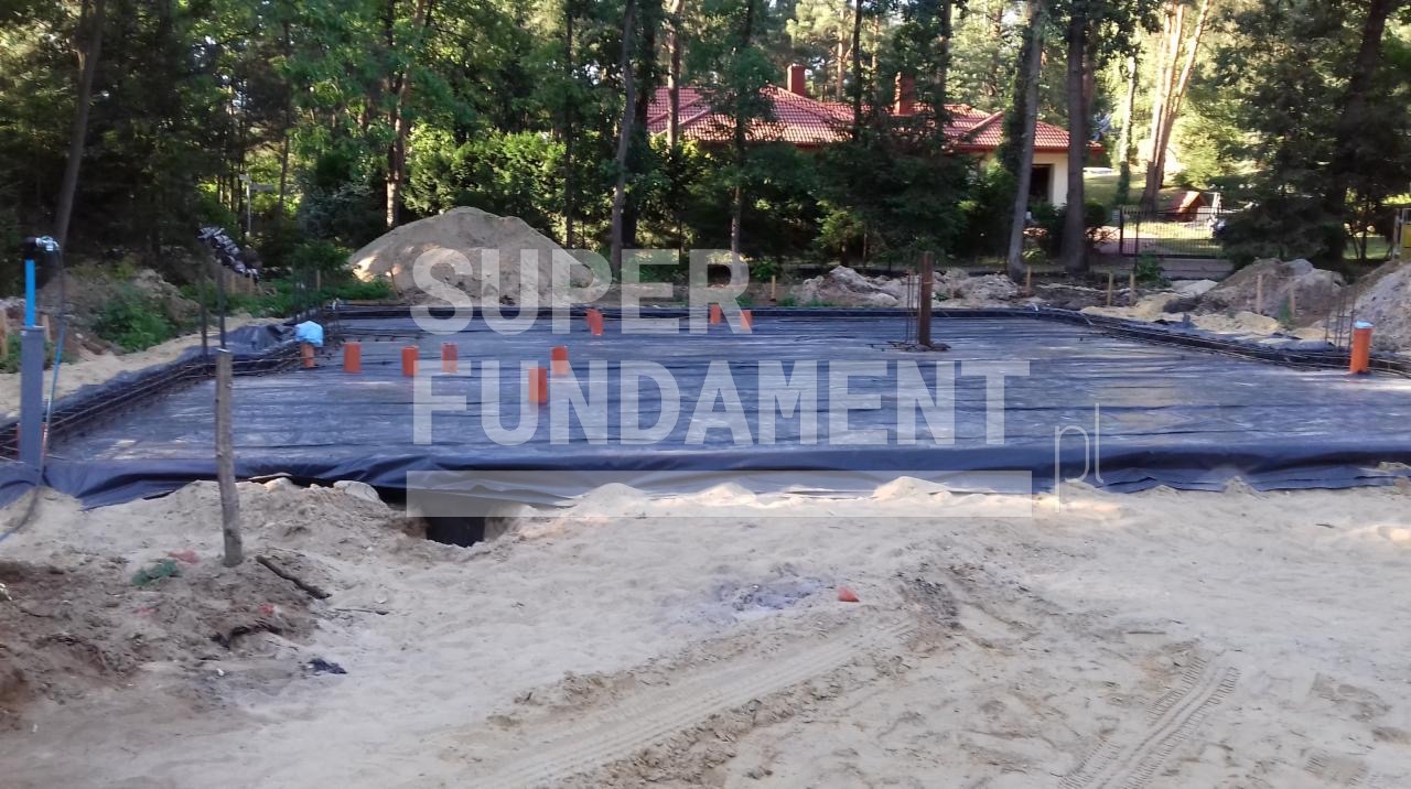 Przygotowany teren pod fundamenty z folią, zbrojeniem i wystającymi rurami wentylacyjnymi na tle lasu i domu z czerwoną dachówką. Częściowe ujęcie.