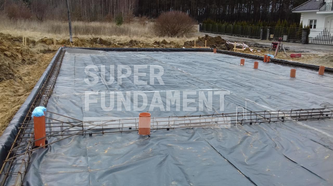 Przygotowany teren pod fundamenty z izolacją, zbrojeniem i pionowymi rurami kanalizacyjnymi. Widoczny teren budowy i częściowo wykończony dom.