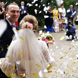 Uśmiechnięty pan młody w granatowym garniturze i fioletową muchą obejmuje pannę młodą w koronkowej sukni z welonem, obsypywanych konfetti na tle wiwatujących gości i fotografa.