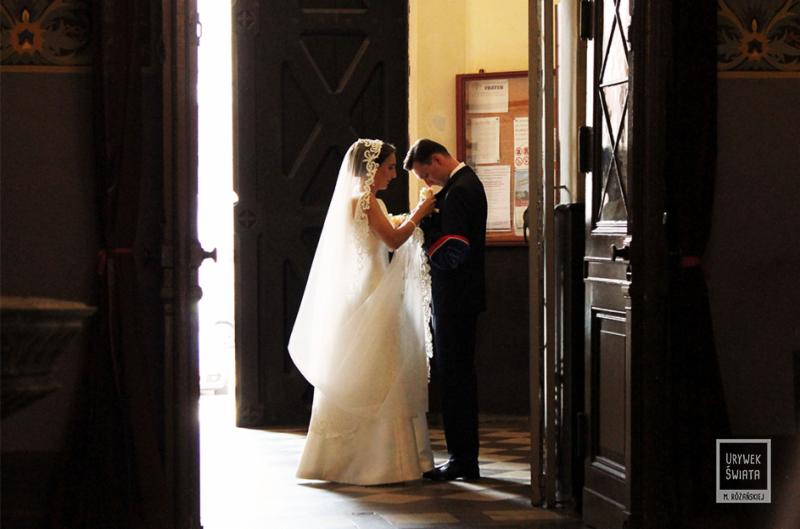 Panna młoda w koronkowej sukni i welonie poprawia butonierkę panu młodemu w ciemnym garniturze przed starymi drewnianymi drzwiami kościoła.