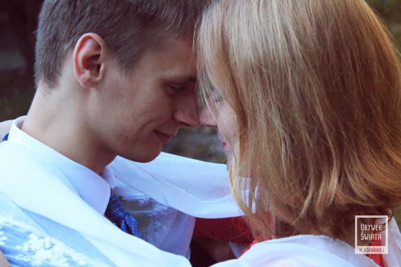 Młoda para w zbliżeniu, czoła dotykają się, owinięci w biało-czerwoną flagę, on w białej koszuli i niebieskim krawacie, ona z włosami w odcieniach blondu.