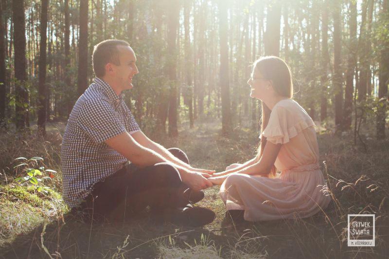 Para siedzi na trawie w lesie, trzymając się za ręce, w tle promienie słoneczne przebijają się przez drzewa.
