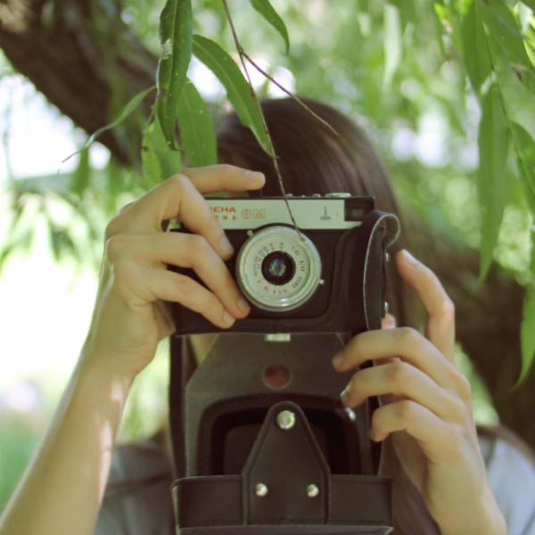 Osoba trzyma w rękach stary aparat fotograficzny marki Chaika, osłonięty skórzanym futerałem, z liśćmi drzew w tle.
