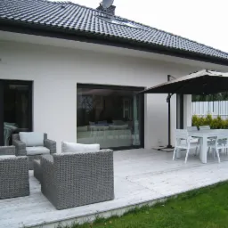 Nowoczesny taras z białym drewnianym podestem, szarymi meblami rattanowymi i białym stołem z krzesłami pod parasolem w ogrodzie przy białym domu z dużymi oknami.