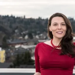 Portret uśmiechniętej kobiety w czerwonej sukience i koralami, na tle miejskiego krajobrazu z zalesionym wzgórzem w tle.