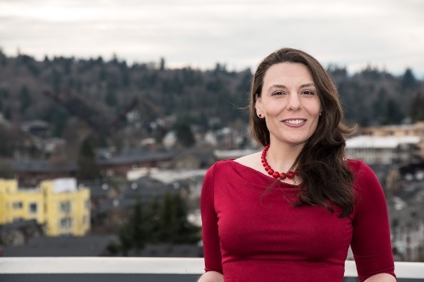 Portret uśmiechniętej kobiety w czerwonej sukience i koralami, na tle miejskiego krajobrazu z zalesionym wzgórzem w tle.