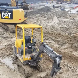 Dwa żółte minikoparki Volvo i CAT w błotnistym terenie budowy; w tle widoczne fundamenty i pracownicy.