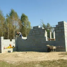 Wznoszenie ścian domu z betonu komórkowego; murarz w trakcie pracy na tle drzew i błękitnego nieba; widoczna taczka i narzędzia.