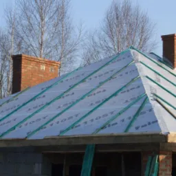 Budowa dachu domu murowanego: membrana dachowa z zielonymi łatami na deskowaniu, widoczne kominy z cegły i fragment konstrukcji więźby dachowej w tle błękitnego nieba.