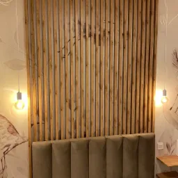 Drewniane lamele na ścianie za wezgłowiem łóżka, z wiszącymi żarówkami na tle ściany z motywem florystycznym.