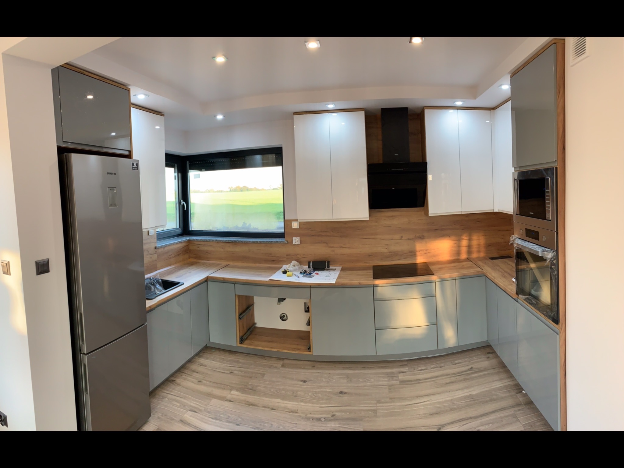 Nowoczesna kuchnia w trakcie montażu: szare szafki dolne, białe górne, drewniany blat i sprzęt AGD, widoczne narzędzia i elementy instalacji.