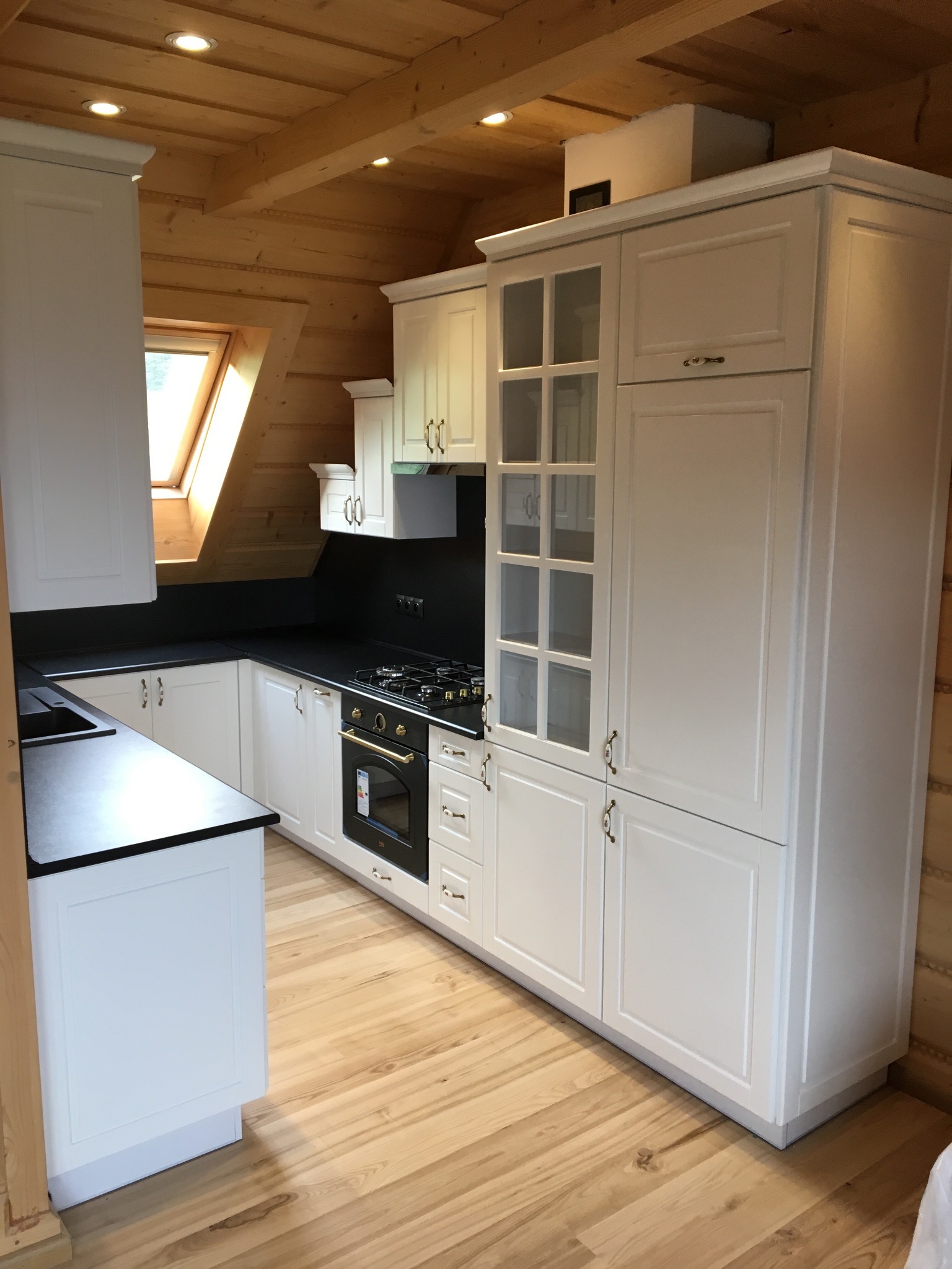 Biała kuchnia w stylu rustykalnym z drewnianymi ścianami i podłogą, czarny blat, okno dachowe, złote wykończenia piekarnika.