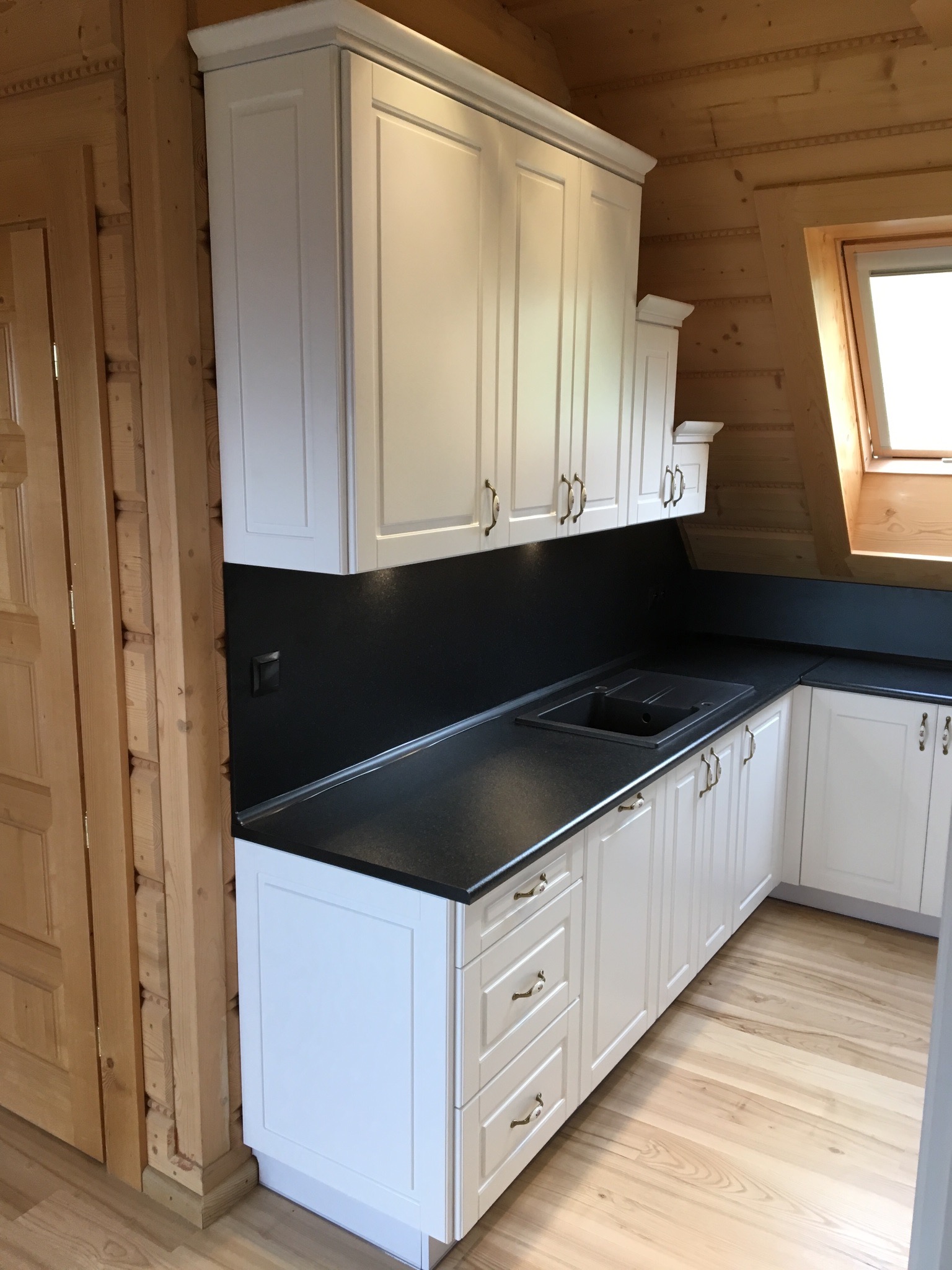 Biała zabudowa kuchenna z czarnym blatem w drewnianym domu ze skośnym oknem dachowym.