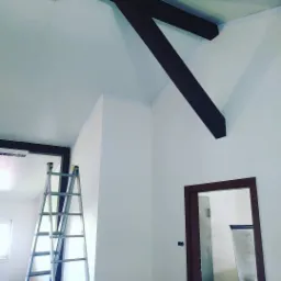 Wnętrze po odświeżeniu, białe ściany i sufit, ciemne drewniane belki stropowe, aluminiowa drabina oparta o ścianę, widoczne wykończenia wokół drzwi i pod sufitem.