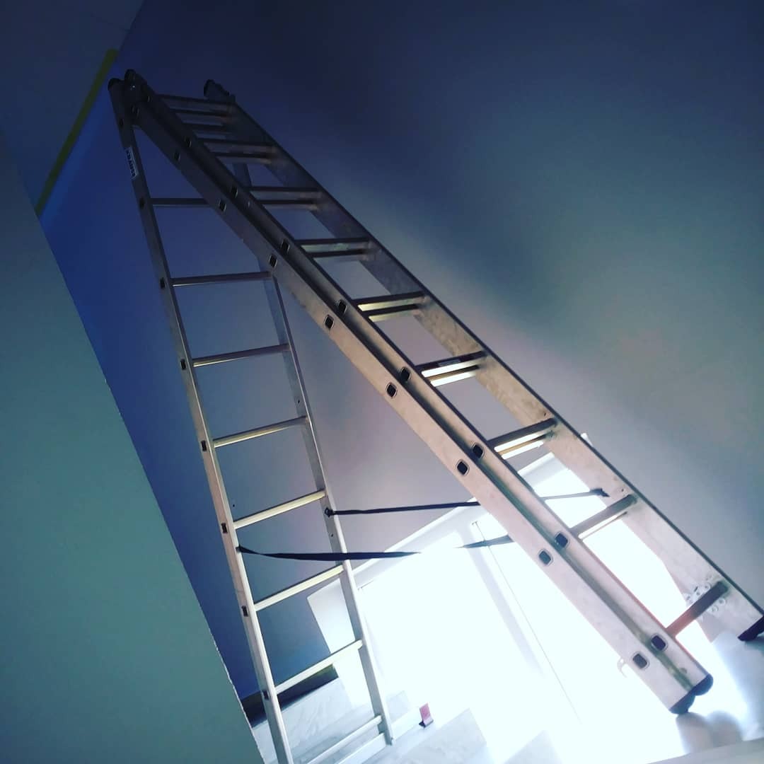 Aluminiowa drabina rozstawiona w pomieszczeniu, widoczne świeżo pomalowane fragmenty ściany w kolorze niebieskim, zabezpieczone taśmą malarską przy suficie.