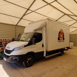 JA-RMUŻ - Biały samochód dostawczy Iveco z logo firmy przeprowadzkowej Yogi, zaparkowany w zadaszonym magazynie. Widoczne palety i konstrukcja dachu.