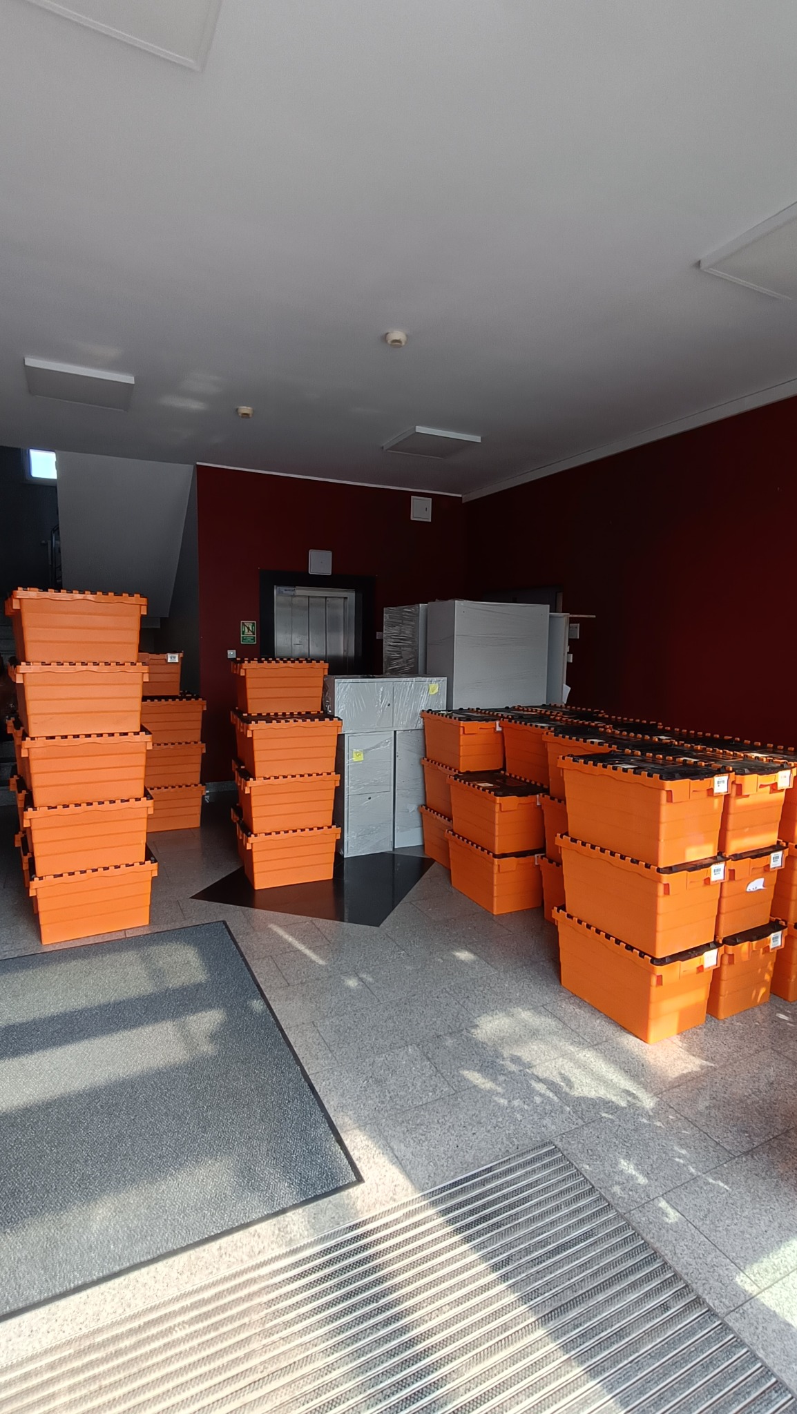 Stosy pomarańczowych plastikowych pojemników do przeprowadzki i zabezpieczone meble w folii stretch w holu budynku z windą.