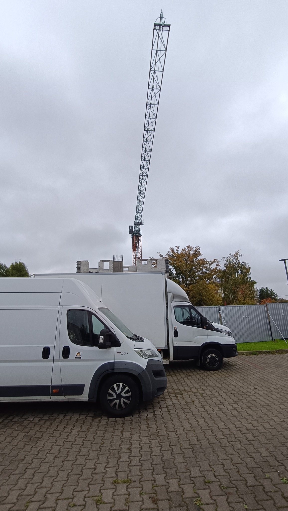 Dwa białe busy transportowe na brukowanym placu, w tle budowa z dźwigiem, pochmurne niebo. Jeden bus marki Fiat Ducato.