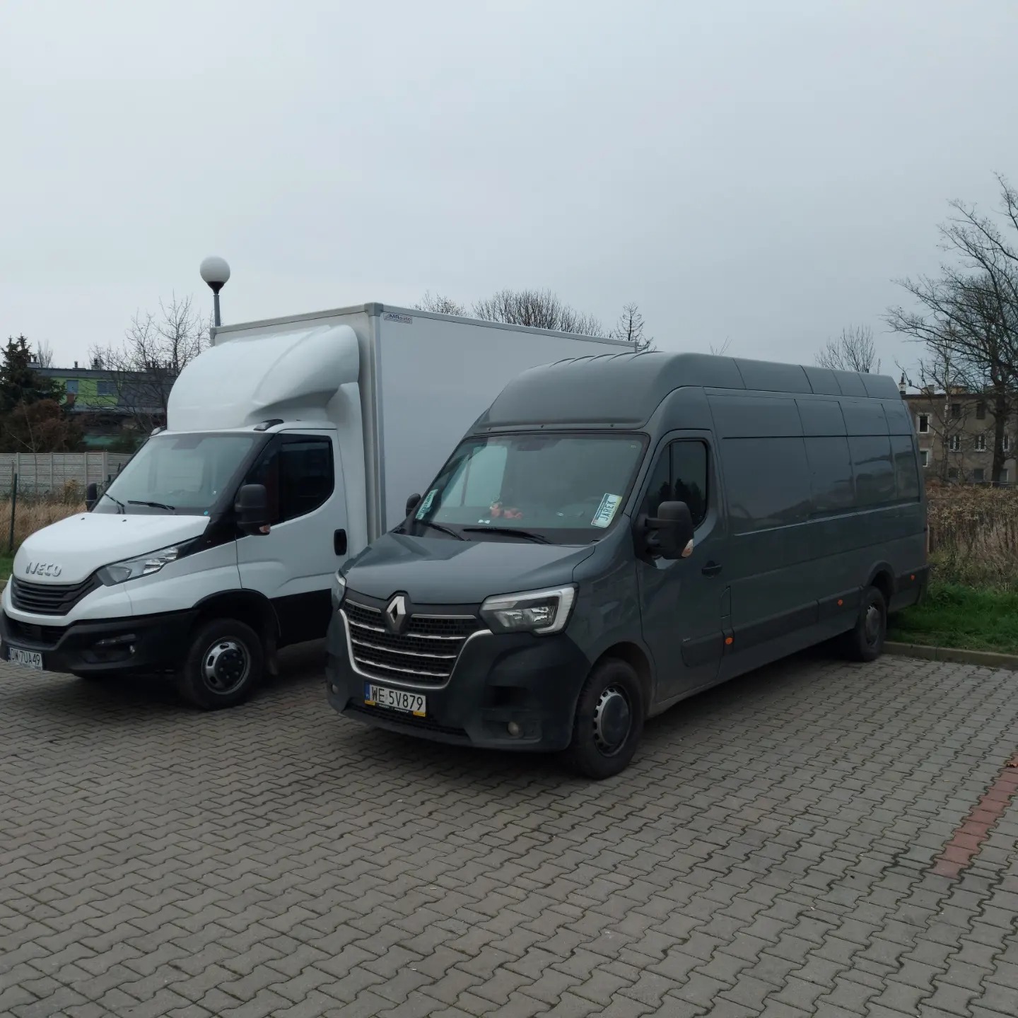 Dwa busy dostawcze: biały Iveco z zabudową kontenerową i szary Renault Master, zaparkowane na brukowanym placu w pochmurny dzień.