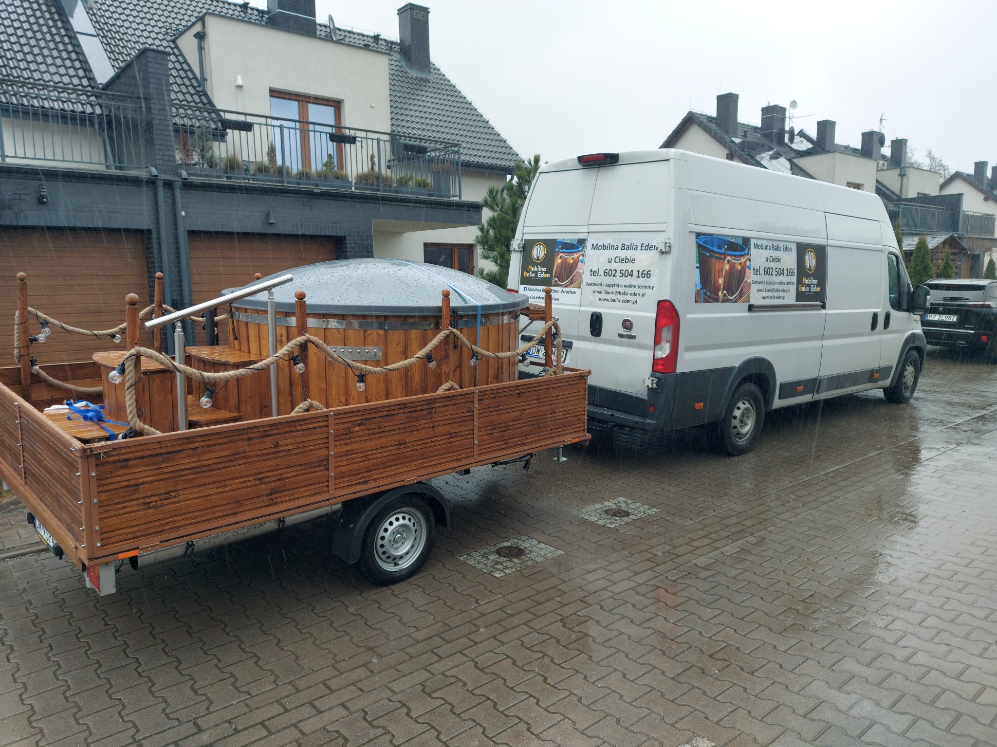 Mobilna balia z hydromasażem, transportowana na przyczepie za busem z reklamą firmy, na tle domów w deszczowy dzień.
