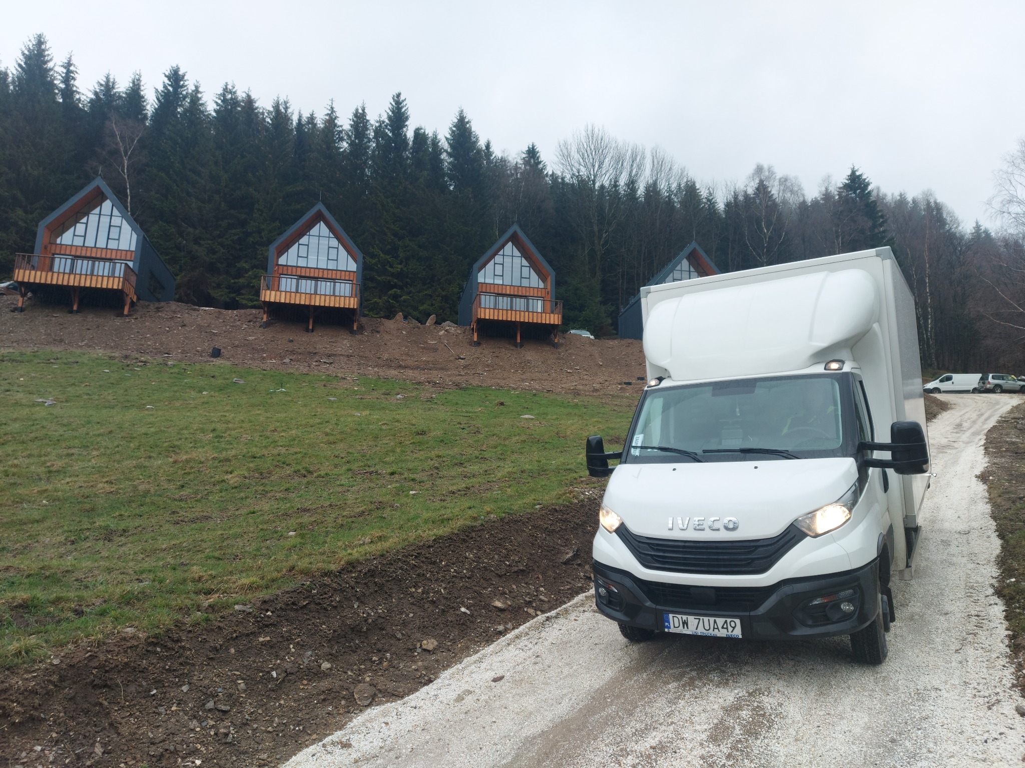 Biały samochód dostawczy Iveco na żwirowej drodze w kierunku nowoczesnych domków letniskowych na wzgórzu, las w tle.