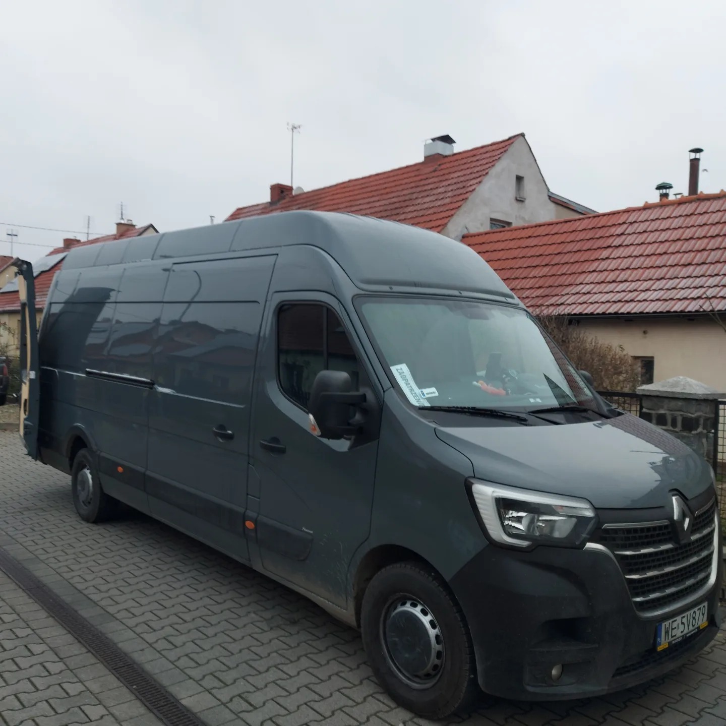 Szary Renault Master zaparkowany na brukowanej ulicy przed domem z czerwoną dachówką w pochmurny dzień.