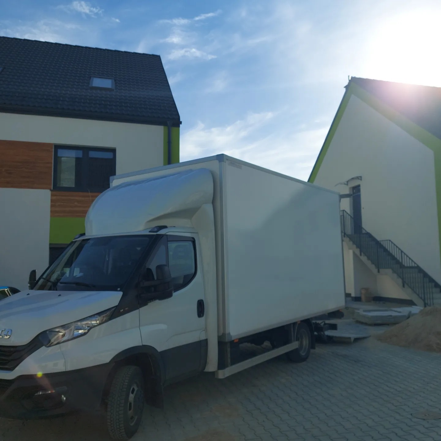 Biały samochód dostawczy Iveco z nadbudową kontenerową na tle nowoczesnych domów jednorodzinnych w słoneczny dzień.