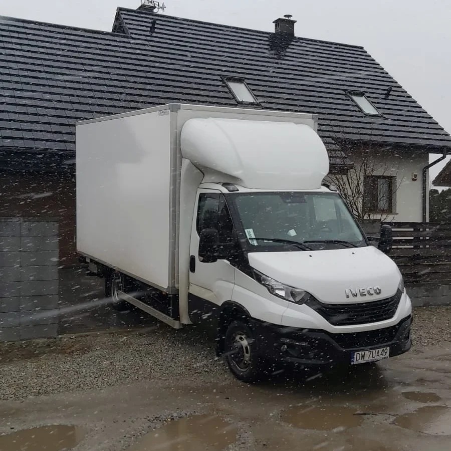 Biały samochód dostawczy Iveco z zabudową kontenerową na tle domu jednorodzinnego podczas opadów śniegu.