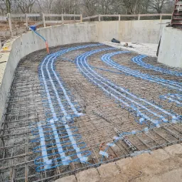 Zbrojenie fundamentu pod budowę z ogrzewaniem podłogowym, widoczne niebieskie rurki grzewcze ułożone w regularnych odstępach na siatce zbrojeniowej, szalunek betonowy w tle.