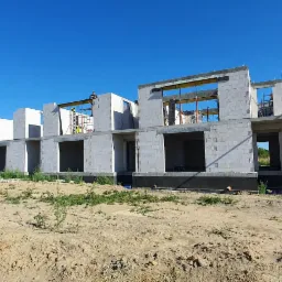 Budowa szeregu domów murowanych z betonu komórkowego, widoczne fundamenty z izolacją, pracownik na rusztowaniu, błękitne niebo.