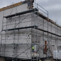 Wznoszenie ścian z białych bloczków na budowie, widoczne rusztowania, fragment zaparkowanego białego busa i zbiornik gazu Linde w tle.