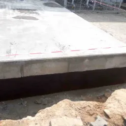 Betonowy fundament z czarną izolacją przeciwwilgociową, częściowo zasypany piaskiem, widoczne wystające pręty zbrojeniowe, na górze mokra powierzchnia betonu, czerwona taśma ostrzegawcza w tle.