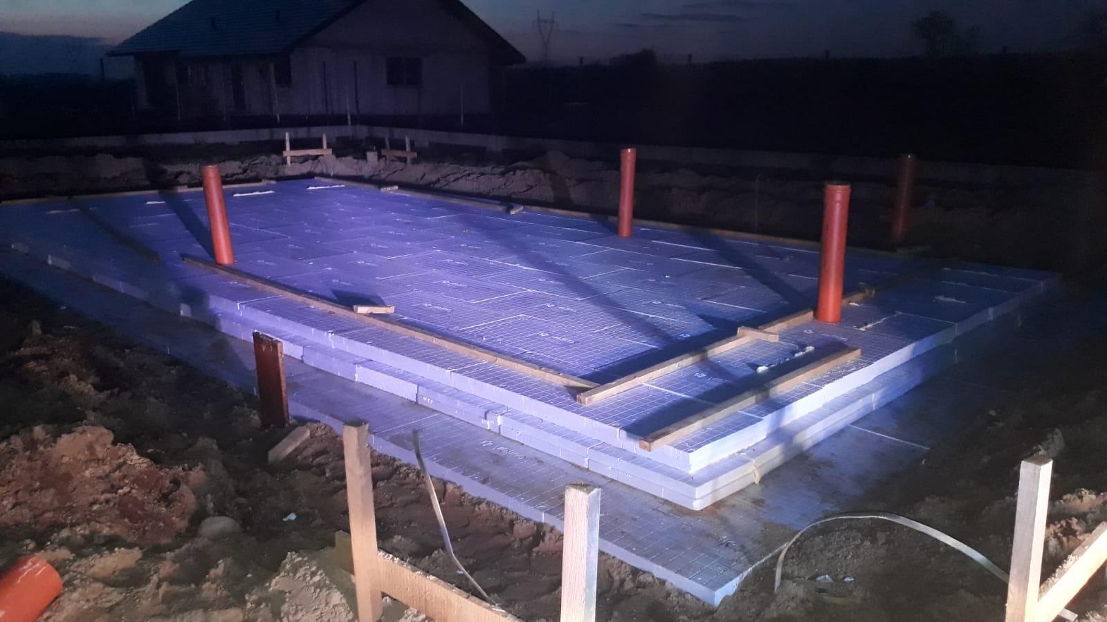 Nocne ujęcie przygotowanego fundamentu z izolacją termiczną i rurami wentylacyjnymi, widoczny fragment domu w tle.