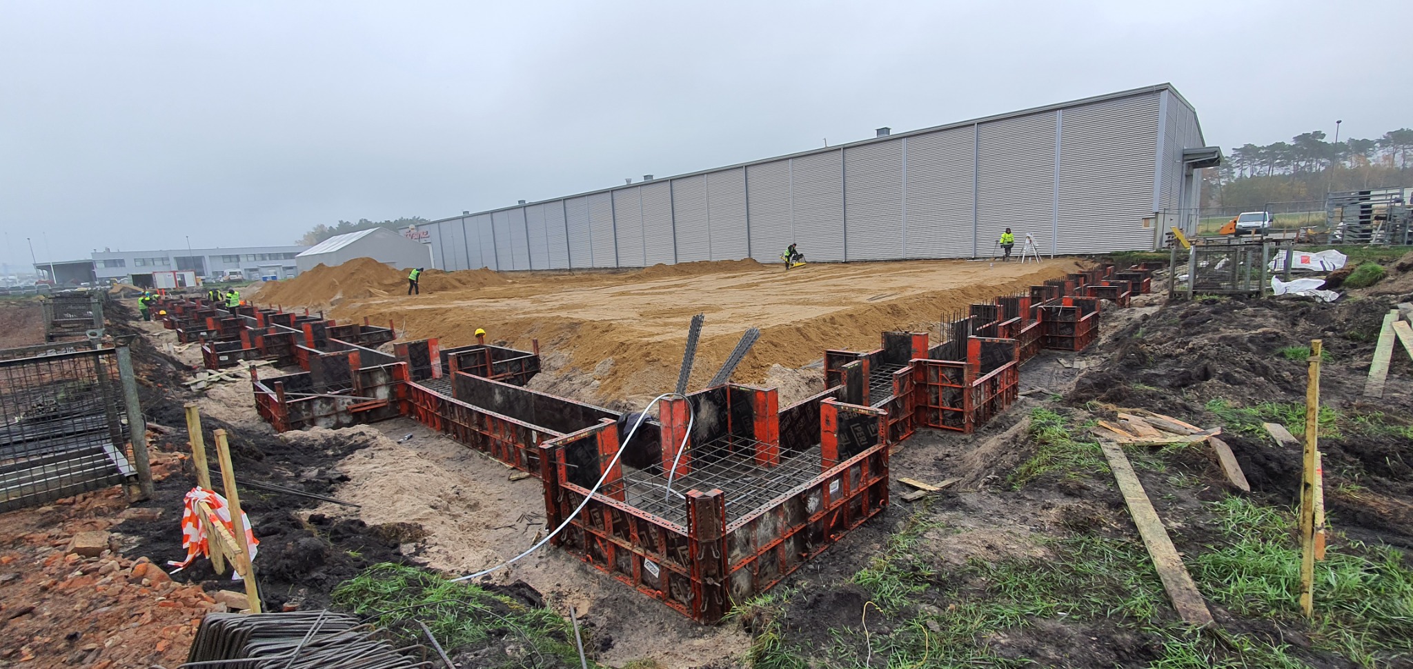 Przygotowanie terenu pod budowę hali: szalunki fundamentowe zbrojone stalą, pracownicy w odblaskowych kamizelkach wyrównujący teren, w tle duży budynek magazynowy.