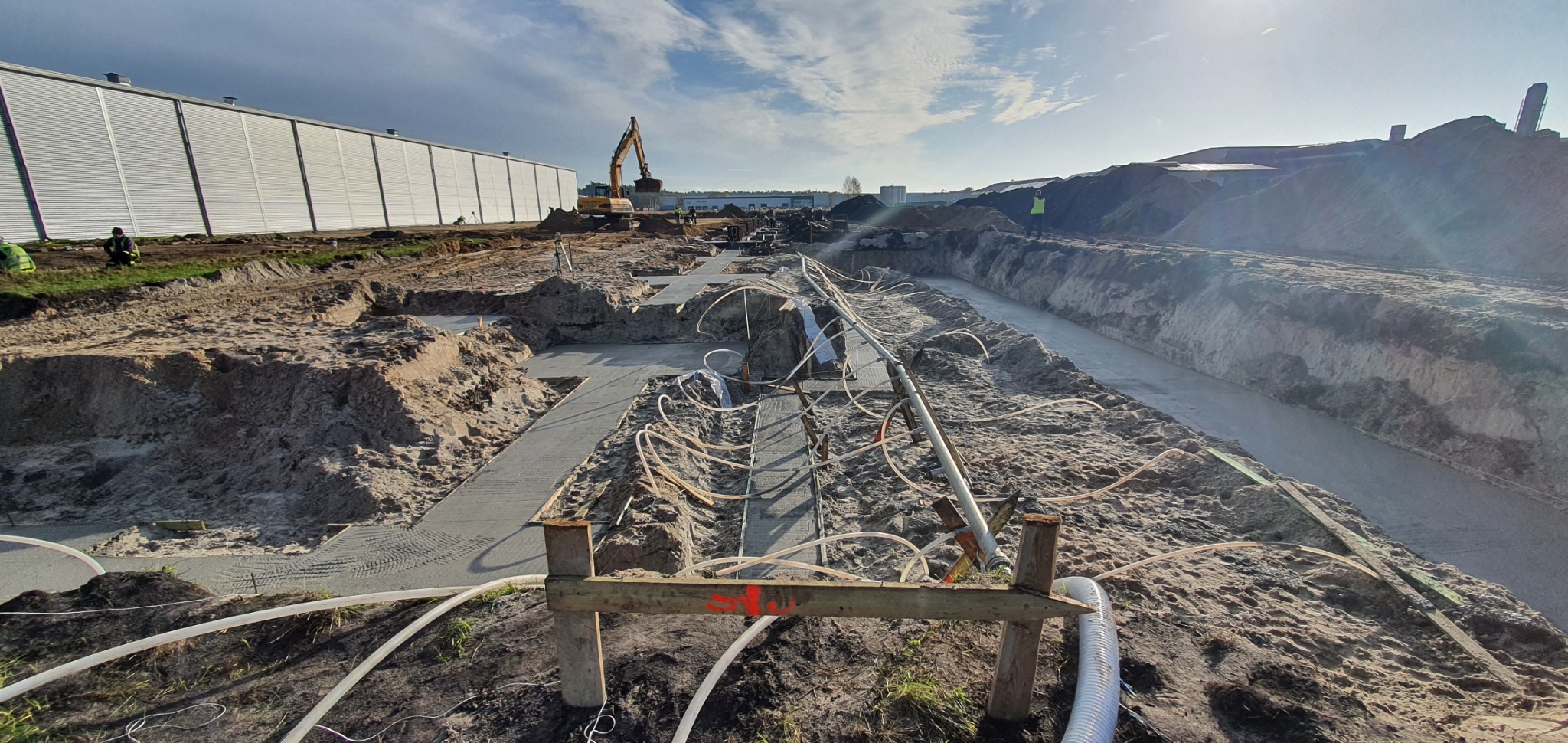 Wykop fundamentów z wylanym betonem, widoczne białe węże do odpowietrzania, w tle budynek przemysłowy i koparka.