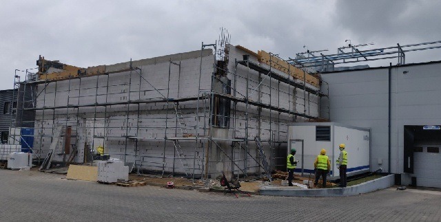 Budowa dwukondygnacyjnego budynku z białych bloczków, widoczne rusztowania, częściowo zamontowane okno, pracownicy w żółtych kamizelkach i kaskach, agregat prądotwórczy obok budynku.