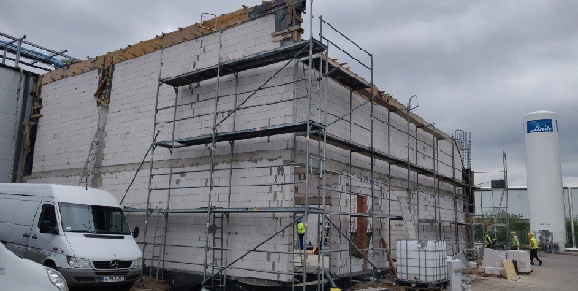Wznoszenie ścian z białych bloczków na budowie, widoczne rusztowania, fragment zaparkowanego białego busa i zbiornik gazu Linde w tle.