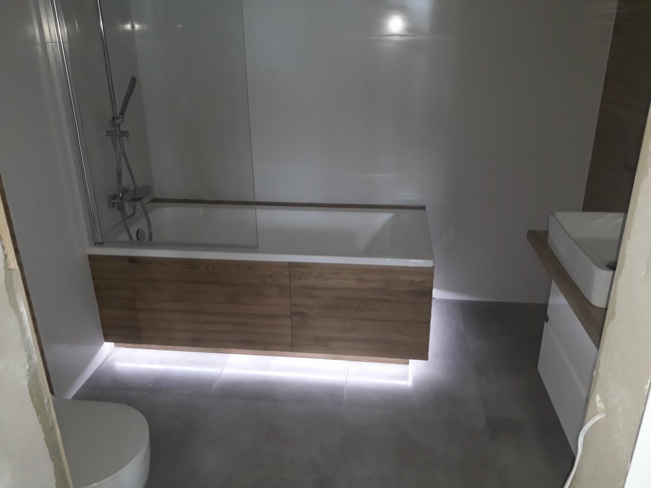 Biała wanna z drewnianą obudową i podświetleniem LED, szklany parawan nawannowy, szare płytki podłogowe, umywalka nablatowa na szafce.