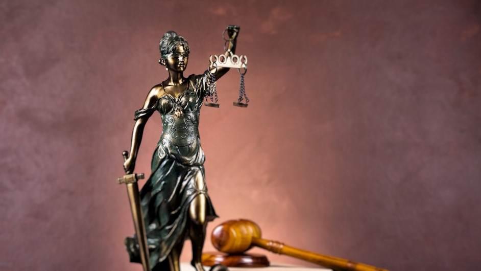 Brązowa statuetka Temidy z mieczem i wagą sprawiedliwości na biurku obok drewnianego młotka sędziowskiego, na tle ściany w odcieniach brązu.