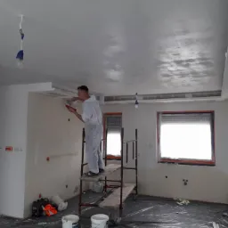 Pracownik w białym kombinezonie na rusztowaniu szpachluje sufit w pomieszczeniu z dwoma oknami, widoczne niedoskonałości sufitu przed finalnym wykończeniem.