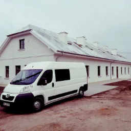 Biały budynek z szarym dachem po renowacji, z zaparkowanym białym busem na pierwszym planie, widoczne świeżo wyrównane podłoże.