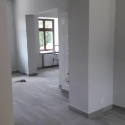 Wnętrze po remoncie: świeżo pomalowane szare ściany, szare panele podłogowe imitujące drewno, widoczne ślady po remoncie i drabina przy oknie.