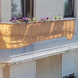 Balkon z metalową balustradą, osłonięty beżową tkaniną, z doniczkami z fioletowymi i białymi kwiatami na górze, widok z dołu na balkon budynku.