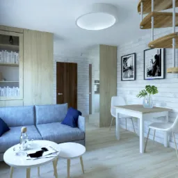 Jasne wnętrze mieszkania z szarą sofą, drewnianymi schodami spiralnymi i białymi meblami, uzupełnione niebieskimi akcentami i czarno-białymi zdjęciami na ceglanej ścianie.