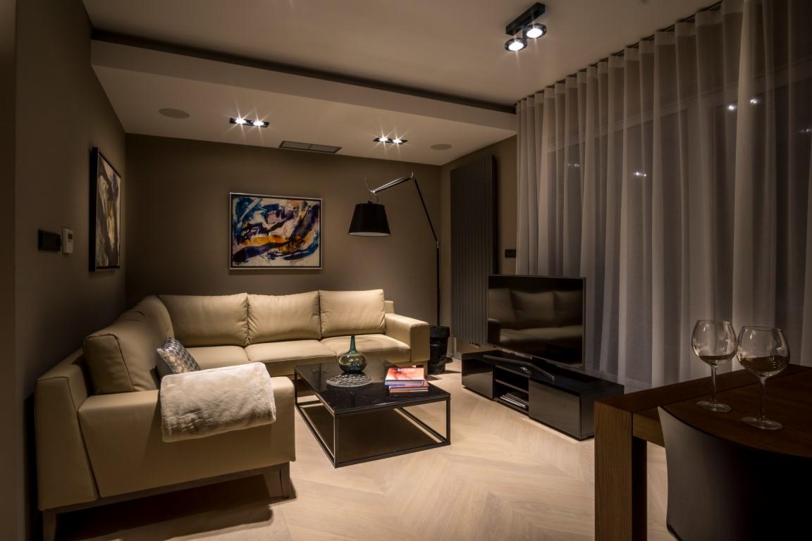 Nowoczesny salon z beżową sofą narożną, czarnym stolikiem kawowym z marmurowym blatem, telewizorem i dwiema lampkami wina na drewnianym stole, oświetlony punktowymi lampami sufitowymi.