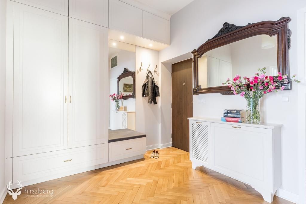 Jasne wnętrze przedpokoju w Będzinie z parkietem w jodełkę, zabudowaną szafą z siedziskiem, dużym lustrem w ozdobnej ramie i białym kredensem z kwiatami.