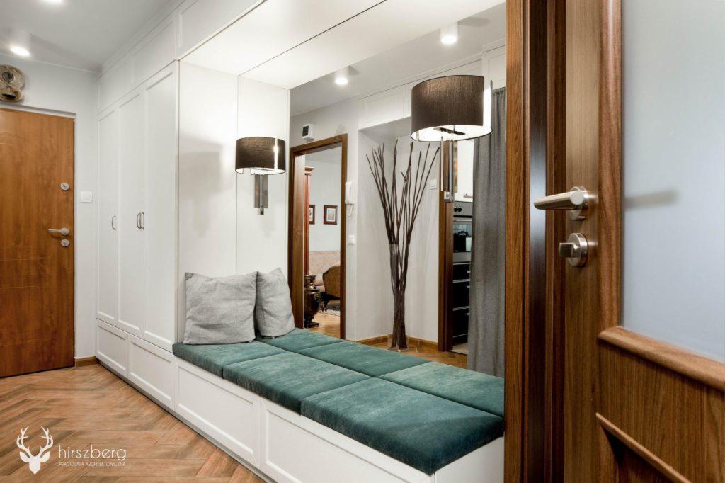 Wnęka z siedziskiem z zielonymi poduchami i szarymi poduszkami na tle białych szaf w przedpokoju z parkietem w jodełkę.