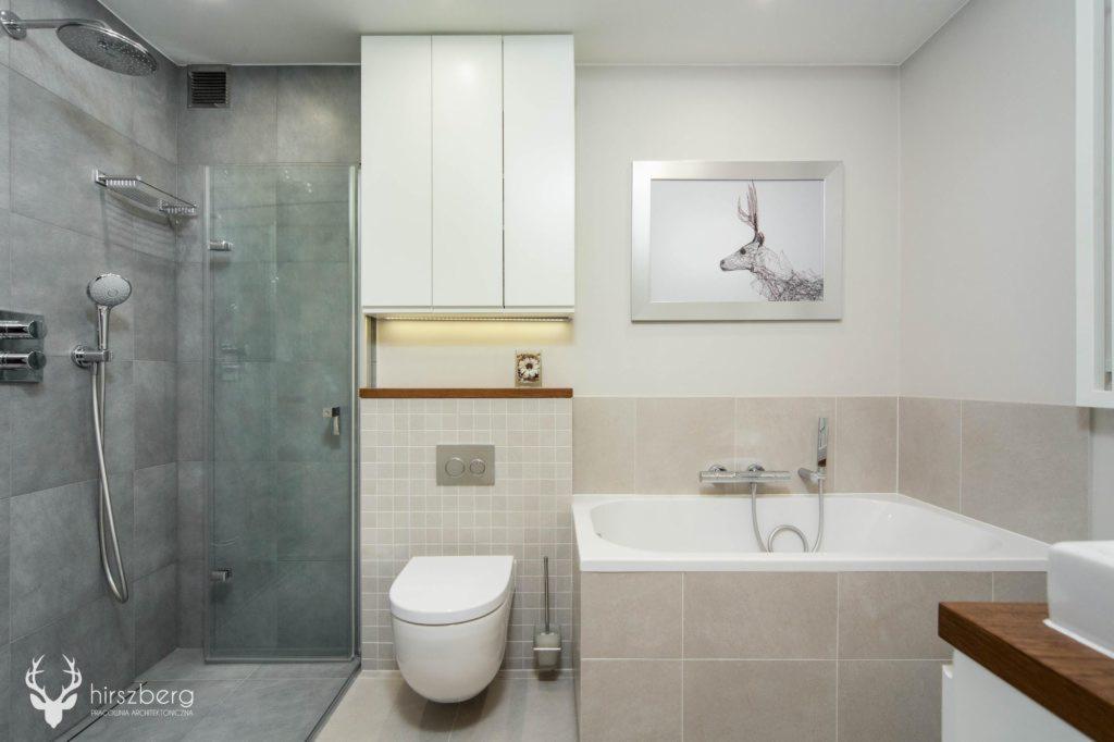 Nowoczesna łazienka z kabiną prysznicową typu walk-in, podwieszaną toaletą, wanną i białymi szafkami wiszącymi, oświetlona listwą LED pod szafkami, z dekoracyjnym obrazkiem jelenia na ścianie.