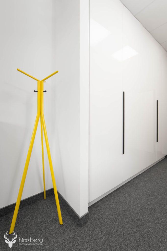 Minimalistyczne wnętrze biurowe z żółtym wieszakiem na ubrania i białymi szafami w zabudowie, szara wykładzina podłogowa, nowoczesny design.