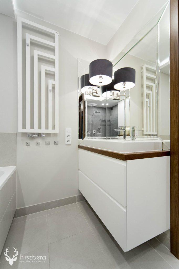 Nowoczesna łazienka w odcieniach szarości i bieli z umywalką wpuszczoną w blat z ciemnego drewna, podwieszana szafka z dwiema szufladami, dekoracyjny grzejnik drabinkowy i lustro z trzema lampami...